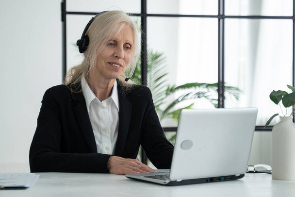 Elderly lady in a headset talks on a video conference, conducts a video conference using a laptop in the office - Фото, изображение