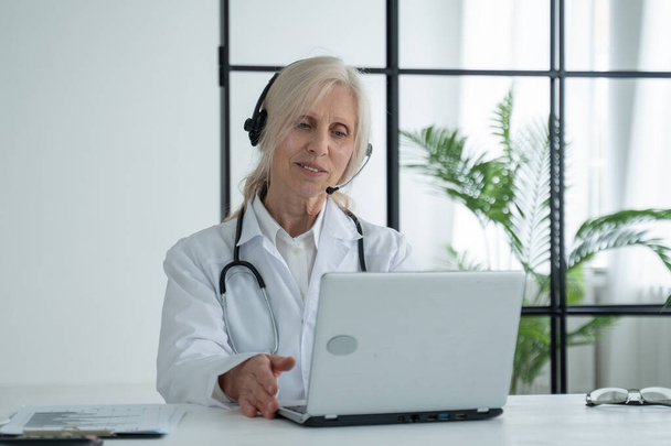 Elderly female doctor with a stethoscope, wearing a lab coat and headset, communicates by video call with a patient via a laptop - Foto, Bild