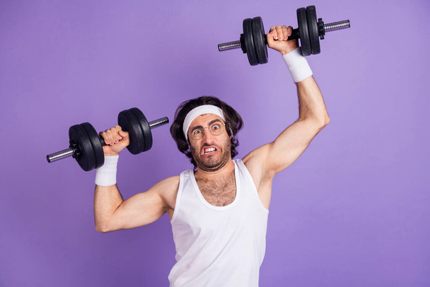 Foto de un joven loco divertido funky en gafas levantando pesadas mancuernas construyendo músculos aislados sobre fondo de color violeta - Foto, imagen