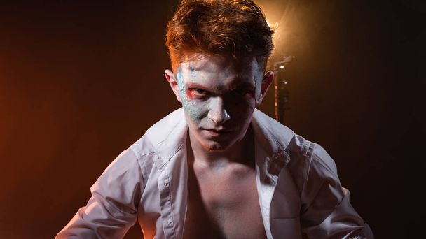 Mad lunatic with a chain on his arm and neck, with makeup on his face against the background of smoke illuminated by yellow light - Photo, image
