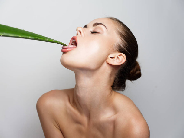 woman with closed eyes and bared shoulders licks an aloe leaf on a light background - Photo, Image