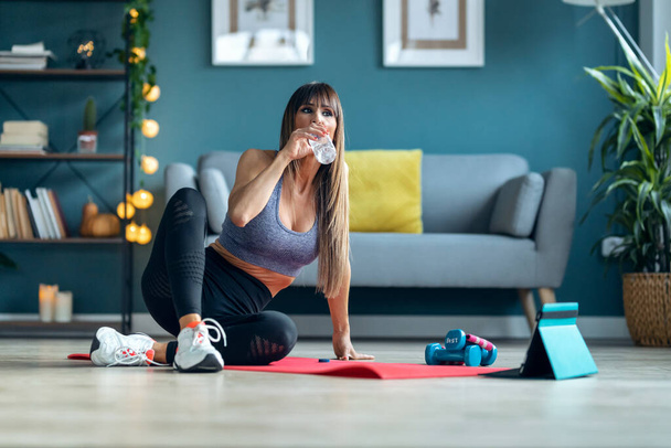 Shot of sporty mature woman drinking water while making a break after online gym classes in the living room at home. - Foto, immagini