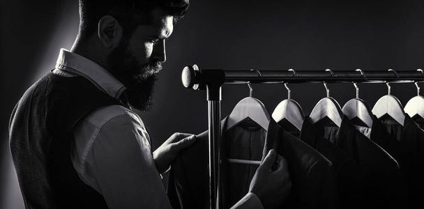 Male suits hanging in a row. Men clothing, boutiques. Tailor, tailoring. Stylish mens suit. Man suit, tailor in his workshop. Handsome bearded fashion man in classical costume suit. Black and white - Photo, image