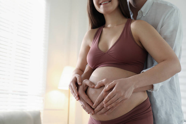 Pregnant young woman making heart with hands on belly and husband near her at home, closeup. Space for text - Photo, image