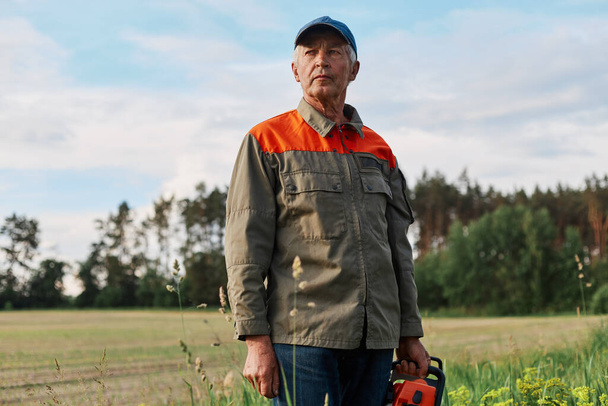 Portrait of mature man wearing jacket and cap posing outdoor in meadow with chainsaw in hands, looking in distance with pensive expression, looks around before working. - Foto, immagini