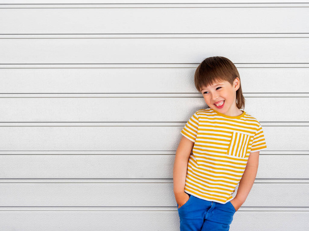 Portrait of smiling boy on white background with stripes. Cute kid stands hands in pockets near light grey wall. Child in yellow t-shirt and blue jeans laughs joyfully. - Fotó, kép