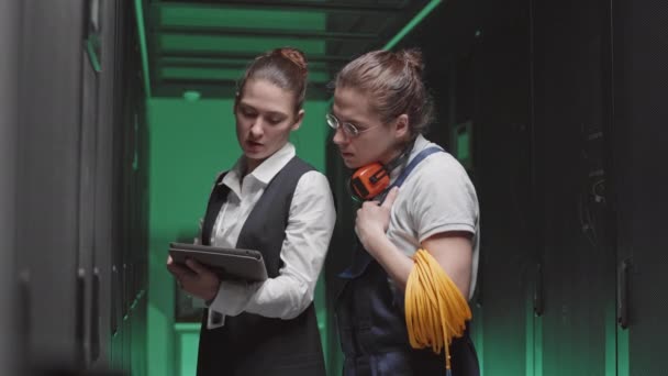 Mittlere Aufnahme einer kaukasischen Ingenieurin, die einen Tablet-Computer in der Hand hält, im Gespräch mit einem jungen männlichen Elektriker mit Drahtgeflechten durch Regalschränke im Serverraum - Filmmaterial, Video