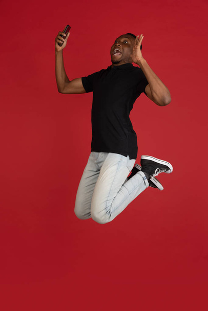 always on mobile. Full length of african young man taking phone while jumping against red studio background. Mobile, motion, movement, business concepts - Photo, Image