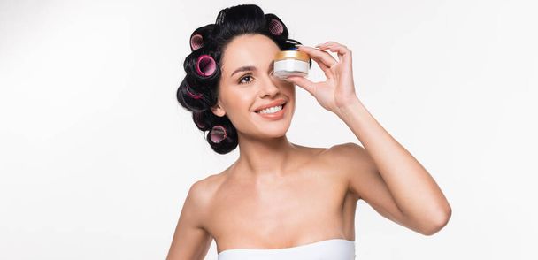 smiling young brunette woman with curlers on hair holding cream container near face isolated on white, banner - Φωτογραφία, εικόνα