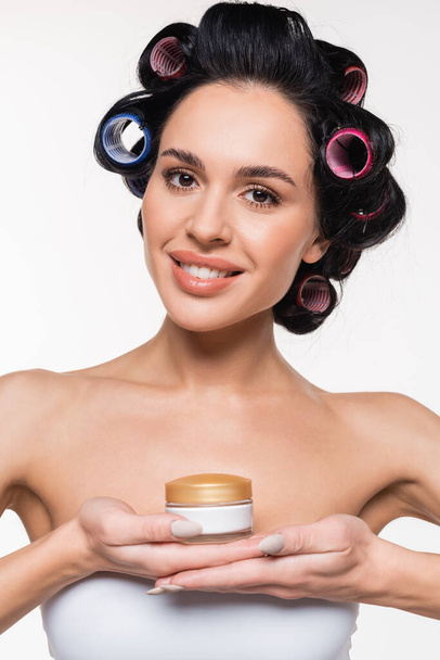 portrait of smiling young woman in white bra with curlers on hair holding cream container in hands isolated on white - Foto, Bild