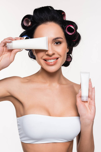 smiling young woman in curlers holding cream tubes in hand and near face isolated on white - Foto, Imagem