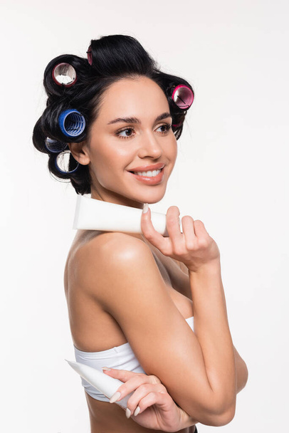 smiling young woman in curlers holding cream tubes isolated on white - Φωτογραφία, εικόνα
