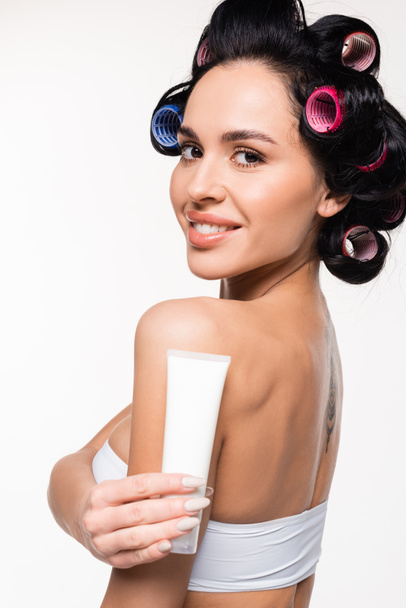 joyeuse jeune femme en bigoudis et top tenant tube crème près de l'épaule isolé sur blanc - Photo, image