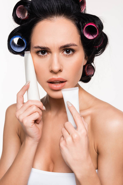 retrato de mujer joven con rizadores en la cabeza sosteniendo tubos de crema cerca de la cara aislada en blanco - Foto, Imagen