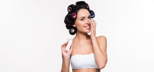 smiling young woman in curlers and bra applying cream on nose and holding tube in hand isolated on white, banner - Fotoğraf, Görsel