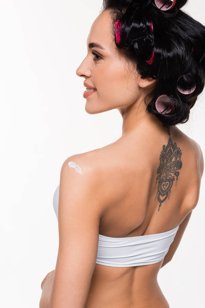 uśmiechnięta młoda kobieta w lokówkach i tatuaż na plecach z kremem na ramieniu odizolowana na biało - Zdjęcie, obraz