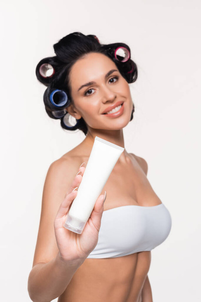 sonriente mujer joven en rulos y sujetador mostrando tubo de crema aislado en blanco - Foto, Imagen