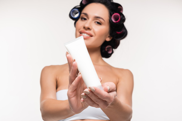 cheerful young woman in curlers and top showing cream tube isolated on white - Φωτογραφία, εικόνα
