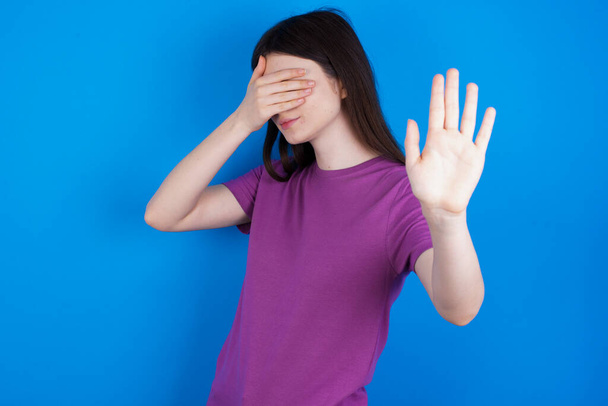 junge Frau bedeckt Augen mit Handfläche und macht Stoppgeste, versucht sich vor allen zu verstecken. - Foto, Bild