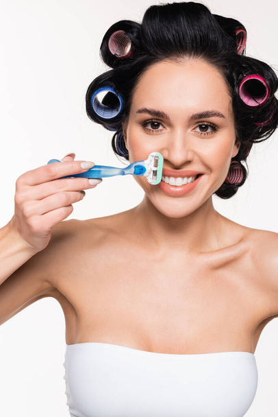 positive young woman in curlers holding razor near face isolated on white - Fotoğraf, Görsel