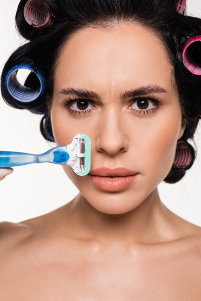 portrait of confused young woman in curlers holding razor near face isolated on white - Fotó, kép