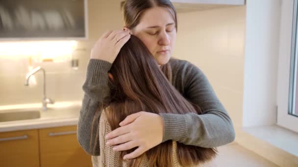 A fiatal anya ölelkezik és vigasztalja feldúlt síró lányát a konyhában. Szülői támogatás és vigasztaló gyermek - Felvétel, videó