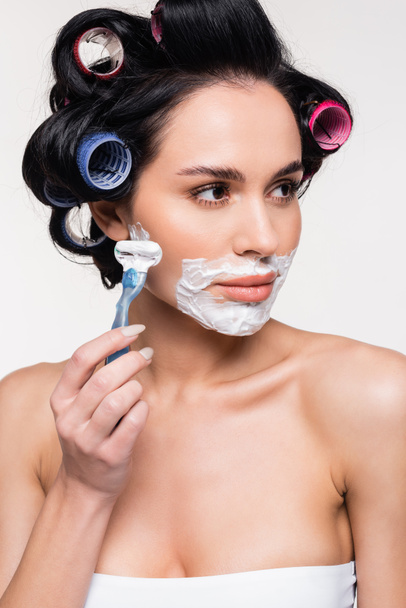 jovem mulher séria em rolos segurando navalha perto do rosto com creme de barbear isolado no branco - Foto, Imagem