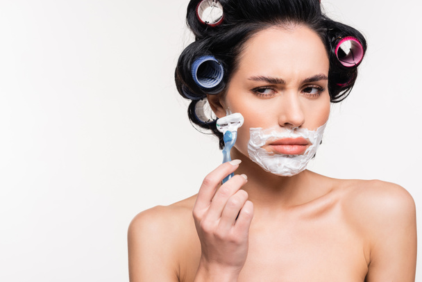 mujer joven avergonzada en rulos con afeitadora cerca de la cara con crema de afeitar aislado en blanco - Foto, Imagen