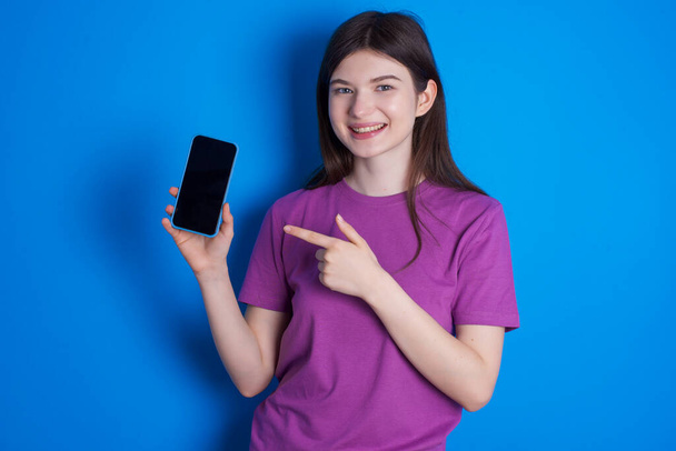 Mujer joven sonriente señalando con el dedo índice en el teléfono móvil con la pantalla vacía en blanco - Foto, Imagen
