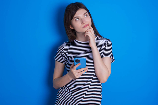 junge Frau denkt tief über etwas nach, benutzt modernes Handy, versucht gute Botschaften zu erfinden, hält Zeigefinger in Lippennähe. - Foto, Bild