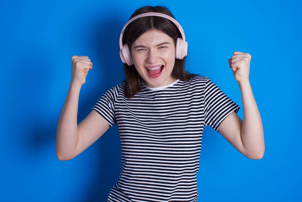 Emotional young woman exclaims loudly feels like winner raises clenched fists keeps mouth opened wears stereo headphones on ears makes yes gesture, listens favourite music - Photo, Image
