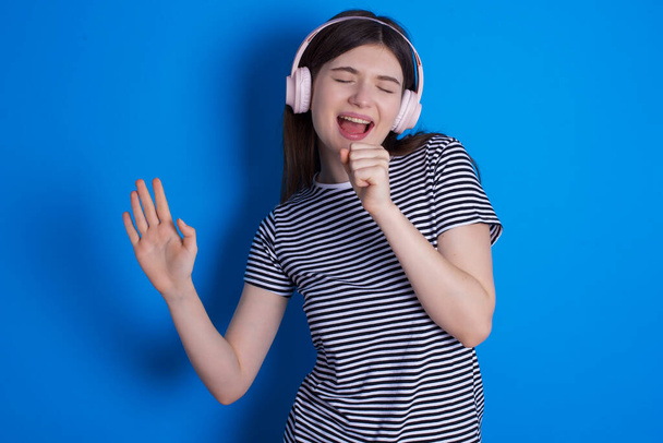 Happy young woman sings favourite song keeps hand near mouth as if microphone wears wireless headphones, listens music - Photo, Image
