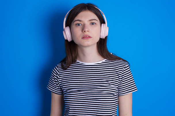 Serious displeased young woman looks puzzled at camera being angry wears stereo headphones listens music while walking at street - 写真・画像