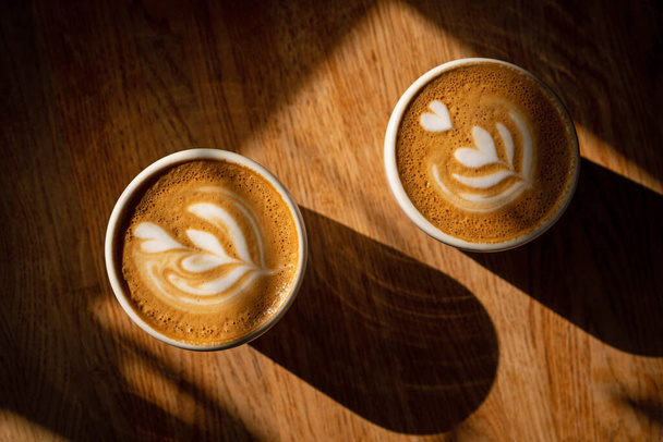 Two cups of fresh cappuccino with latte art on brown wooden table and long deep shadows. Coffee break in cafe. Delicious coffee with milk.  - Photo, Image