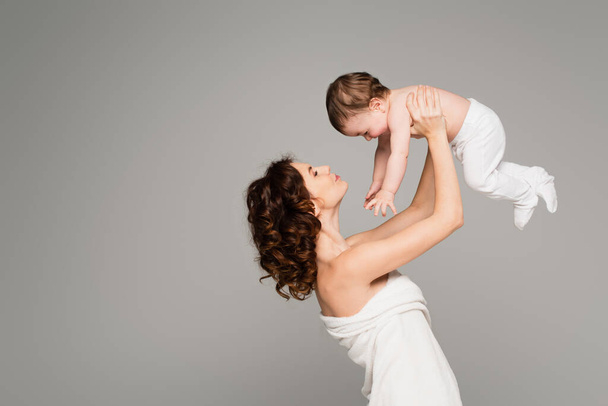 happy mother with bare shoulders holding in arms boy in baby tights isolated on grey - Foto, Bild