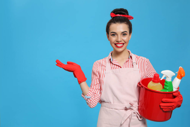 Young housewife holding bucket with cleaning supplies on light blue background, space for text - Fotografie, Obrázek