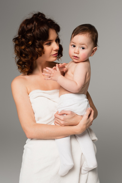 curly mother wrapped in towel holding baby boy in arms isolated on grey  - Foto, afbeelding