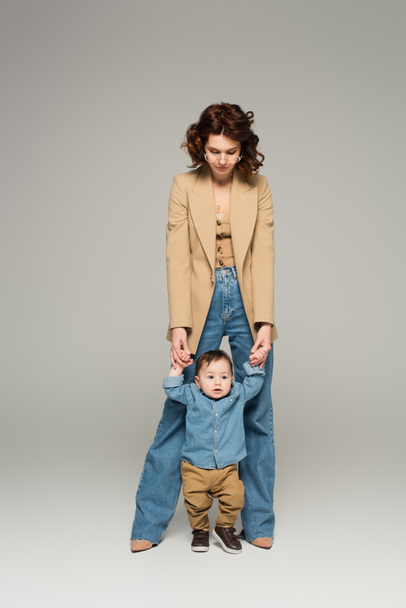 full length of curly mother in glasses supporting and holding hands of baby boy on grey - Фото, изображение