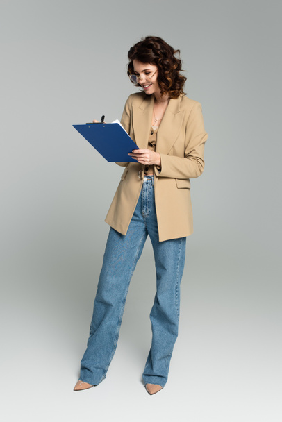 full length of happy businesswoman in glasses and blazer holding clipboard on grey - Foto, Imagen