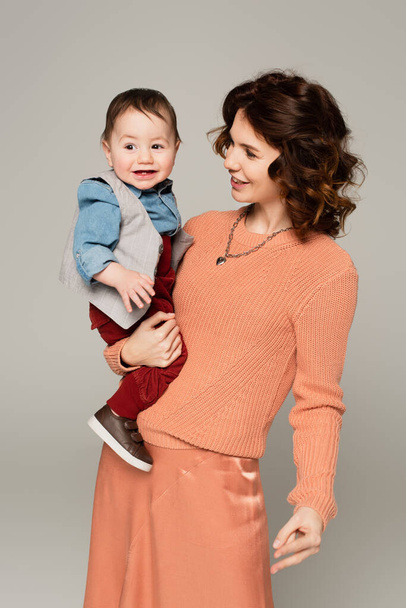 happy woman in sweater holding in arms toddler son isolated on grey - Foto, Imagen
