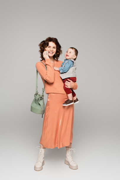 pełna długość szczęśliwy kobieta w satynowy spódnica gospodarstwa w ramiona maluch syn i rozmowy na komórkę na szary - Zdjęcie, obraz