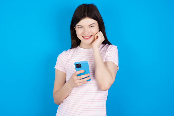 Joyous young brunette woman poses with mobile phone device, types text message on modern smartphone, watches funny video during free time, enjoys good internet connection, - Photo, Image