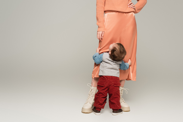 stylová žena v saténové sukni stojící s rukou na boku v blízkosti batole syn na šedé - Fotografie, Obrázek