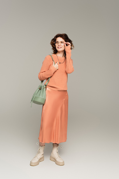 full length of stylish woman in sweater and satin skirt holding leather strap of bag and adjusting glasses on grey  - 写真・画像