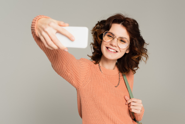 pozitivní žena v brýlích přičemž selfie na smartphone izolované na šedé  - Fotografie, Obrázek