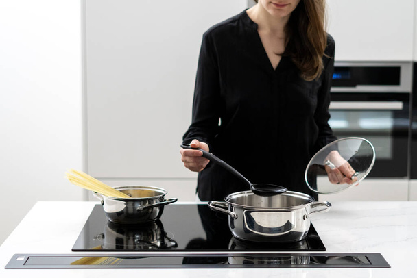Cropped shot of woman boiling water in metal pot to cook spaghetti, holding glass lid and big spoon while cooking on black ceramic induction cooktop in white minimalistic kitchen - Photo, image