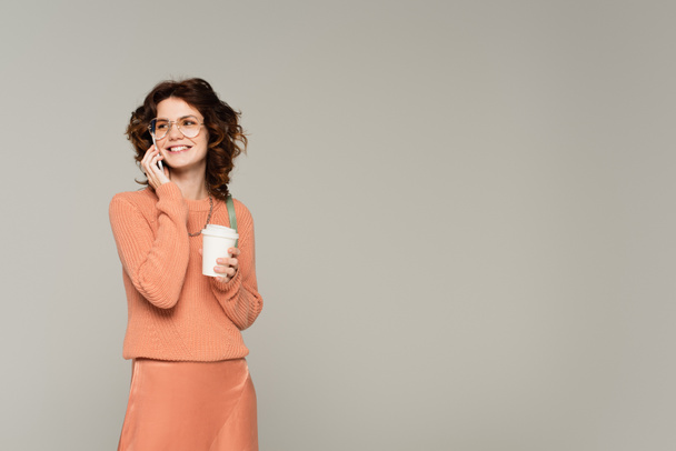 happy woman in sweater holding coffee to go while talking on cellphone isolated on grey  - Foto, Bild