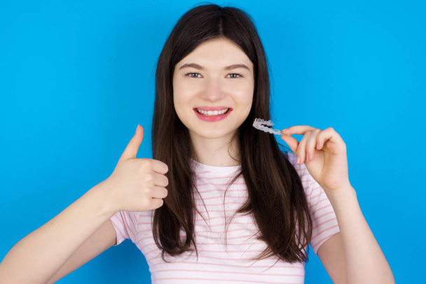 若いブルネットの女性は目に見えないブレースアライナーを保持し、親指を上げて、この新しい治療を推奨します。歯科医療の概念. - 写真・画像