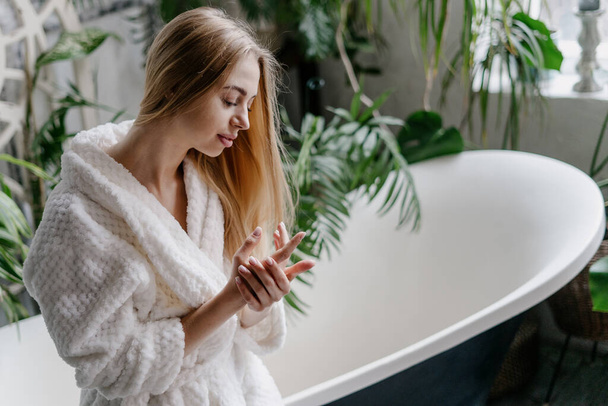 Concept of home spa and natural skincare cosmetics. Smiling and dreamy young woman in white soft bathrobe standing near freestanding bath in cozy bathroom, applying hand cream on skin - Valokuva, kuva
