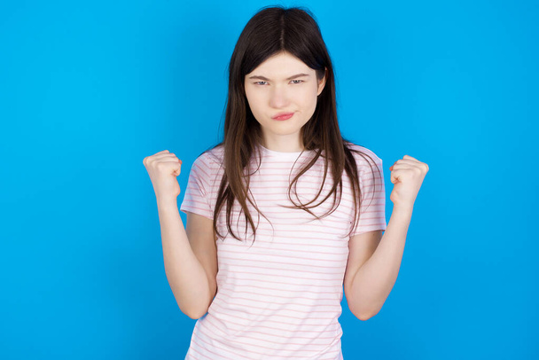 Irritated young brunette woman blows cheeks with anger and raises clenched fists expresses rage and aggressive emotions. Furious model - 写真・画像